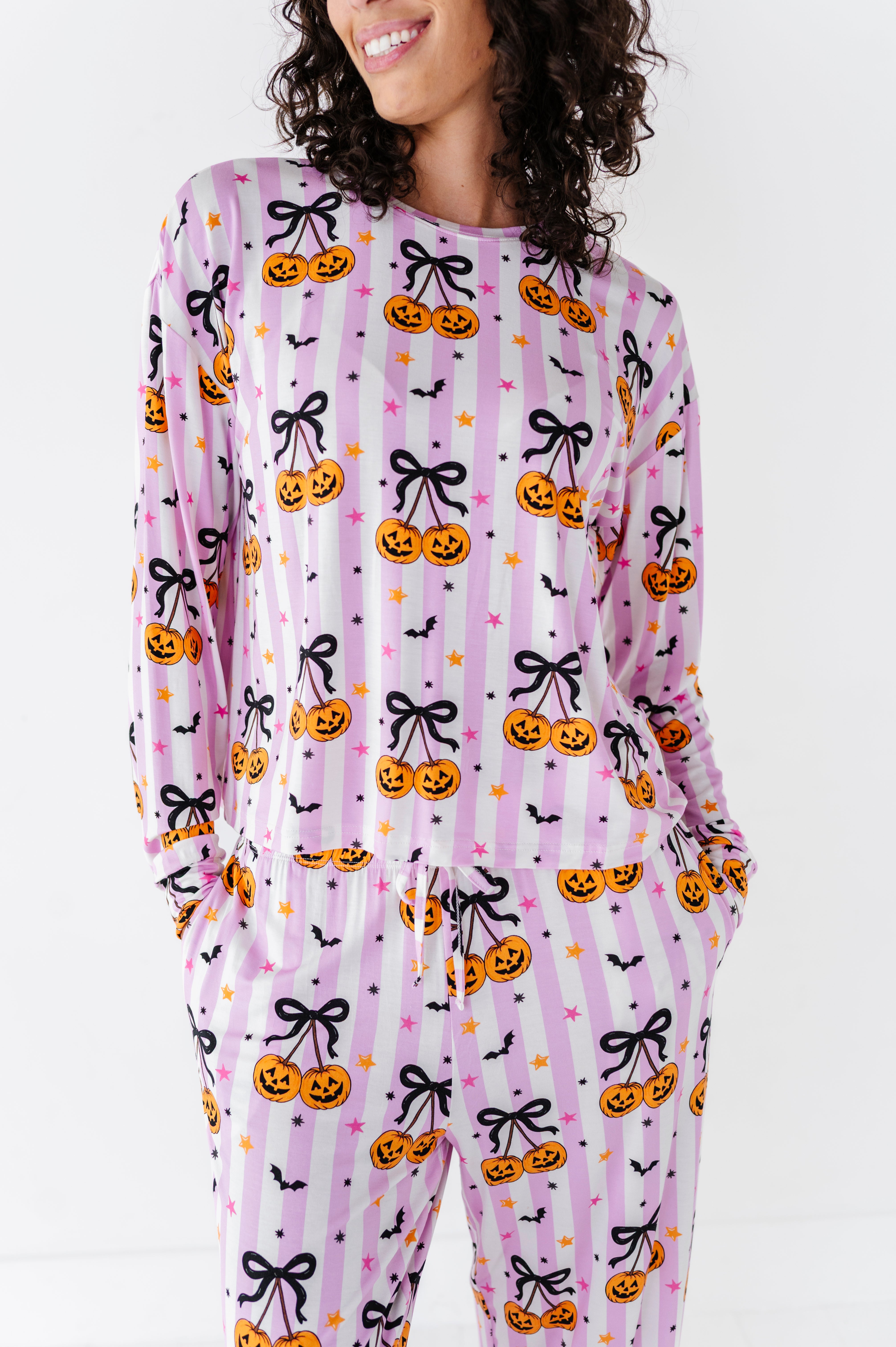 A woman wearing a long-sleeve pajama top and matching pants with a pink and white striped Halloween print featuring pumpkins, black bows, bats, and stars.